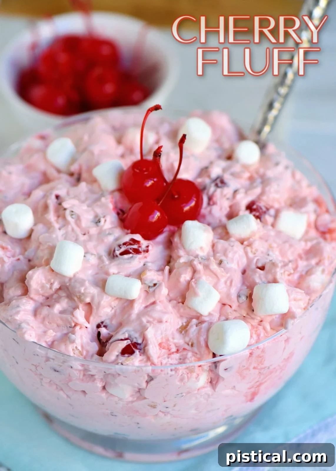 A vibrant clear bowl of Cherry Fluff dessert salad with a serving spoon, garnished with whipped cream and cherries, set against a festive background.