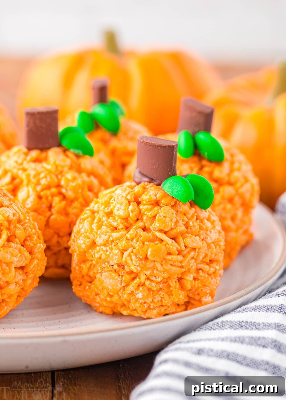 Five or six pumpkin-shaped Rice Krispie treats with chocolate chunk stems and M&M leaves, neatly arranged on a round white plate with a striped napkin beside them.