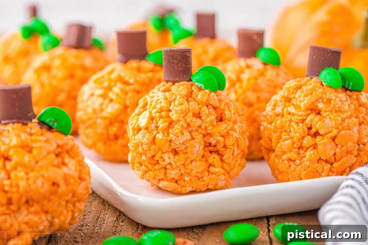 About 10 pumpkin-shaped Rice Krispie treats, each adorned with a chocolate chunk stem and green M&M candy leaves, arranged invitingly on a white platter.