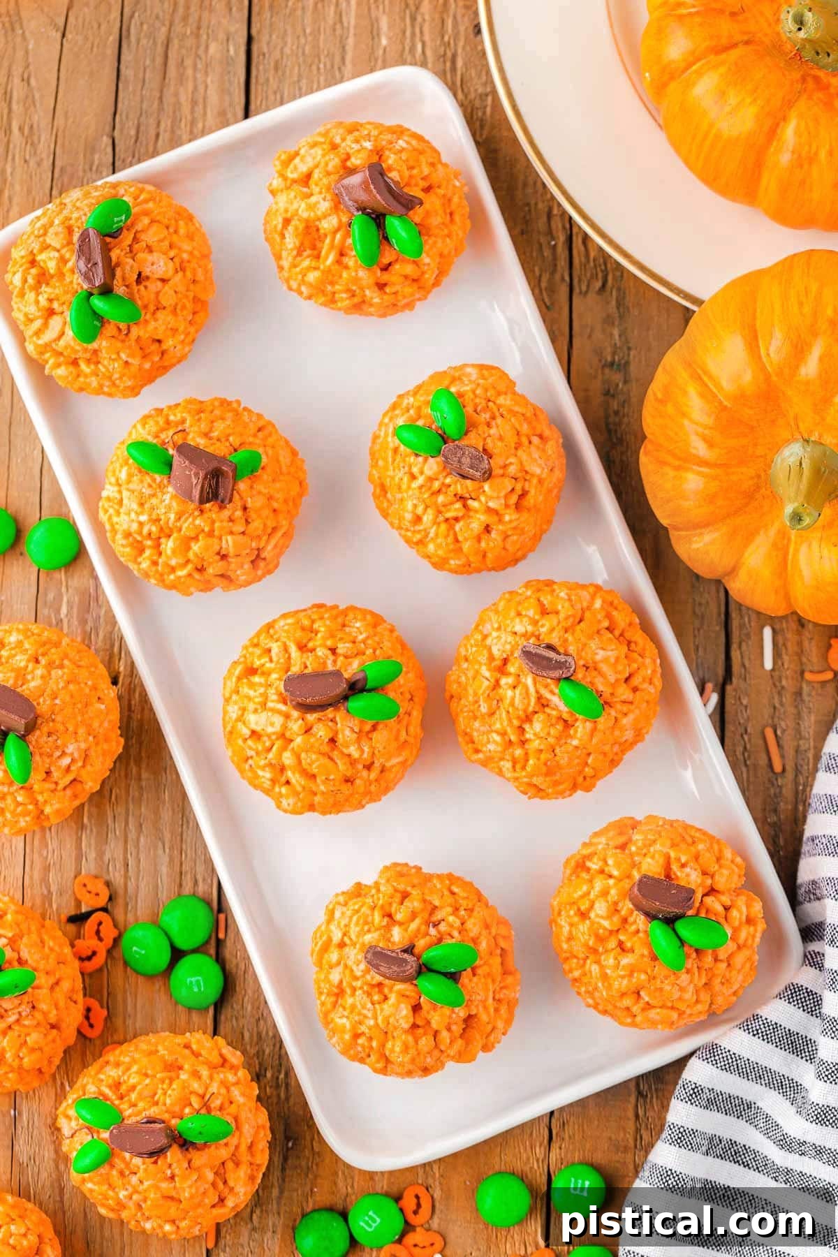 A top-down view of several pumpkin-shaped Rice Krispie treats arranged on a large rectangular white platter, accompanied by decorative mini pumpkins.