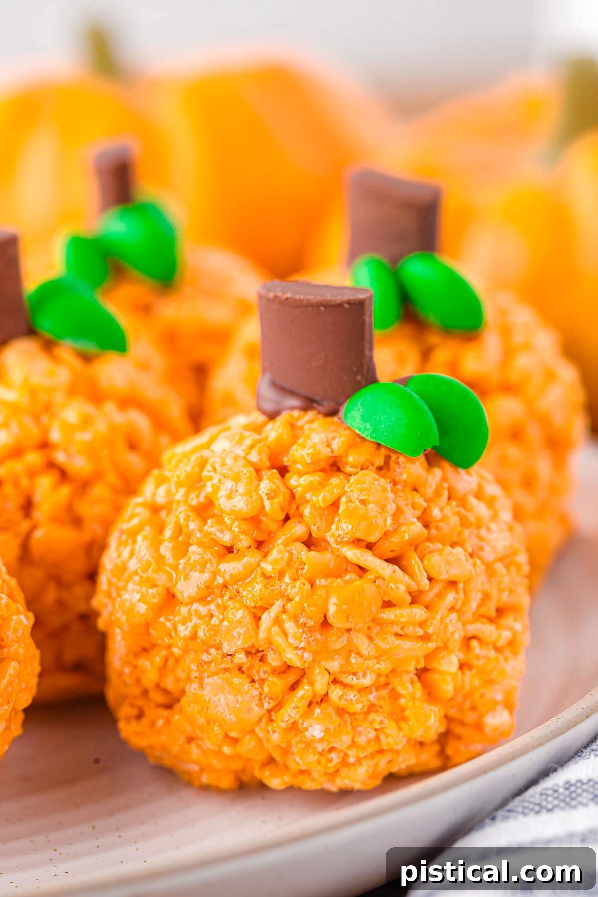 Five or six pumpkin-shaped Rice Krispie treats, some with chocolate stems and green leaves, resting on a round white plate with a striped napkin.