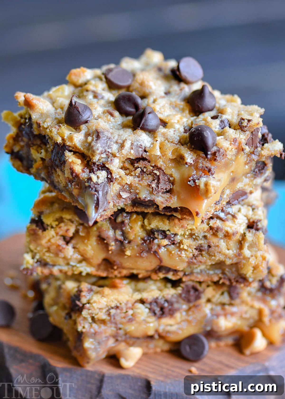 Three decadent cookie bars stacked on a rustic wood board. The bars feature visible layers of peanut butter cookie, gooey caramel, crunchy toffee, and melty chocolate chips, all with a hint of oats in the base.