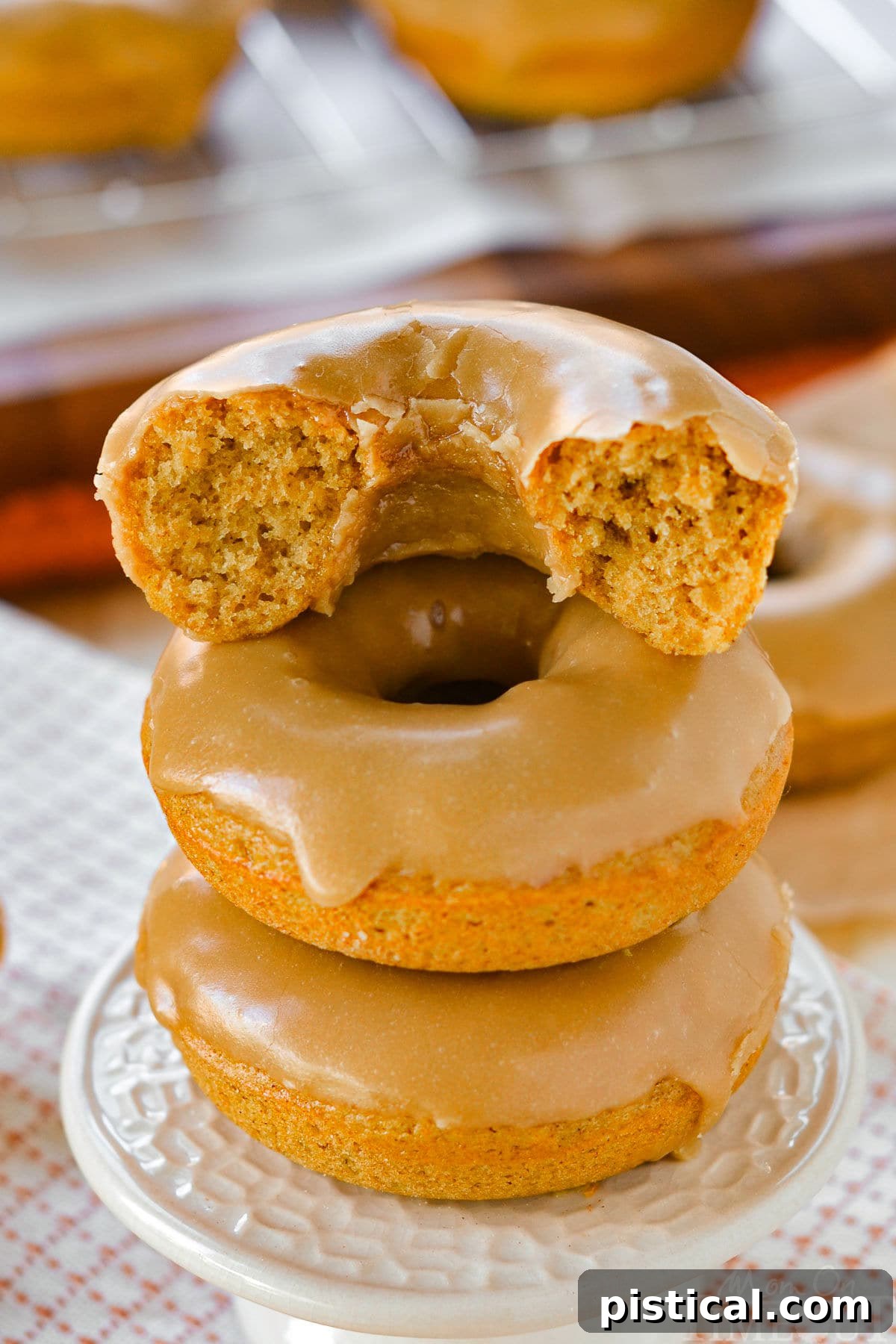 Three perfectly baked pumpkin donuts, glistening with maple glaze, stacked elegantly on a cupcake pedestal. A bite is taken from the top donut, revealing its moist, spiced interior.