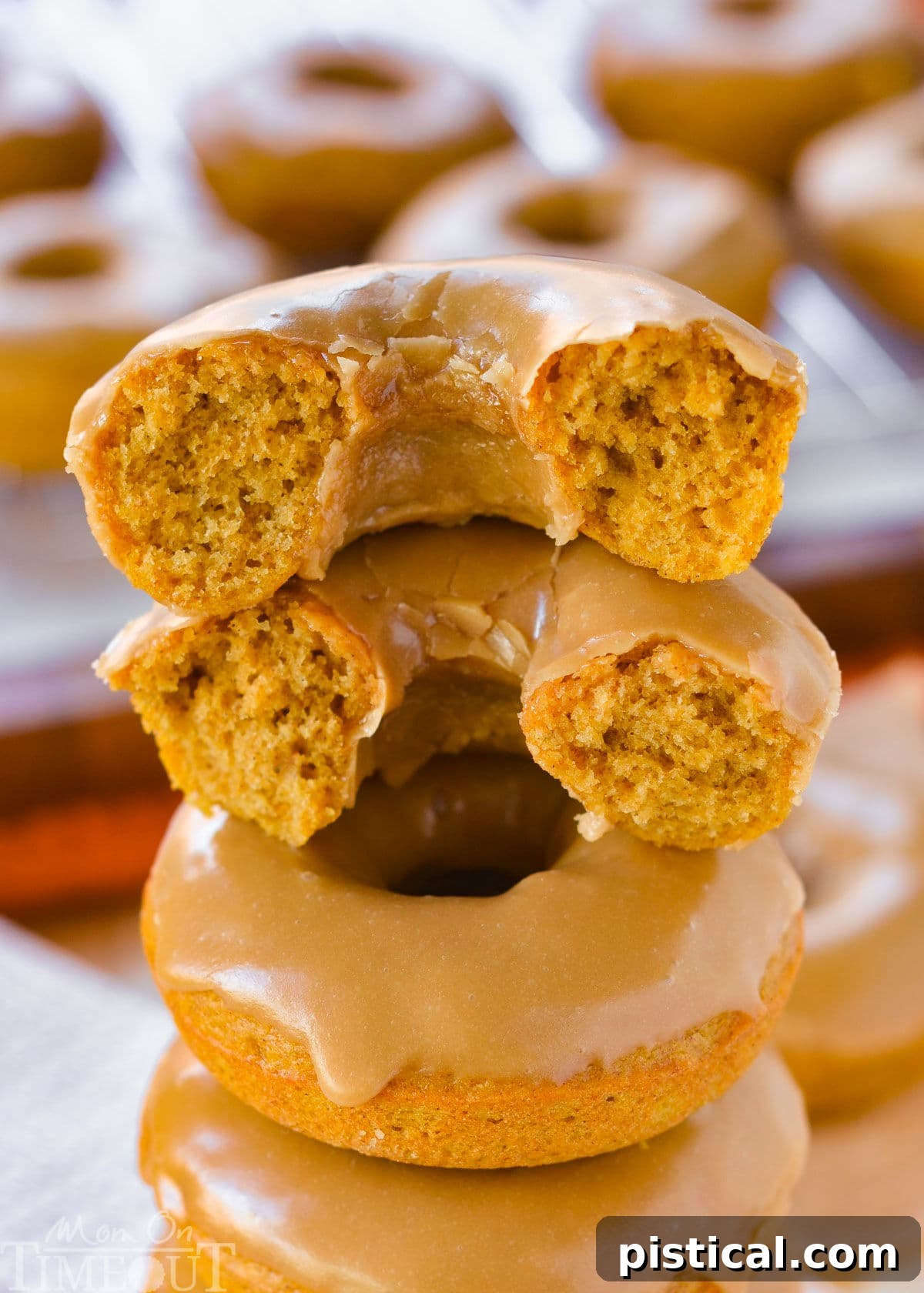 A stack of golden baked pumpkin donuts, with the top donut torn in half to reveal its incredibly moist, cake-like interior and the thick, glossy maple glaze.