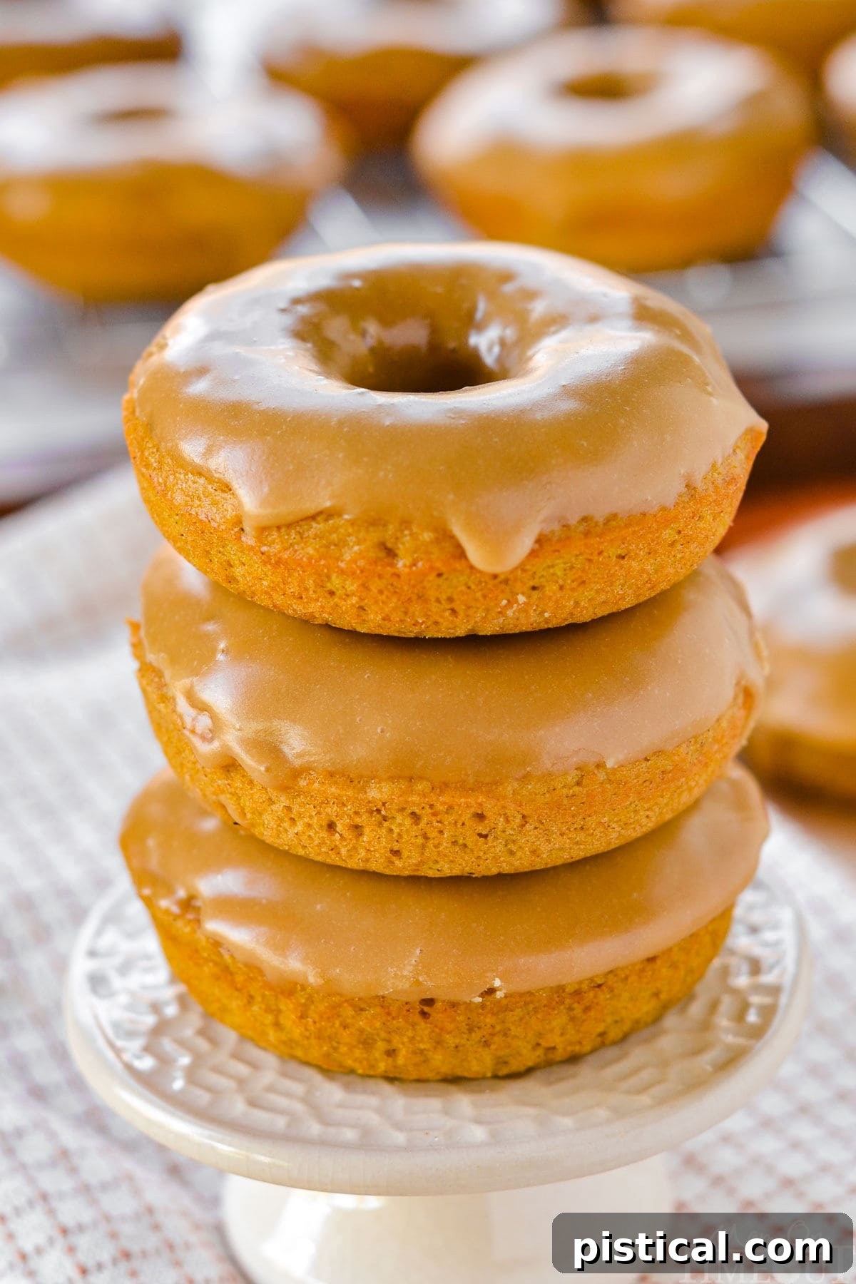 A front-facing view of three beautifully baked pumpkin donuts, stacked on a charming cupcake stand, with their maple glaze glistening.