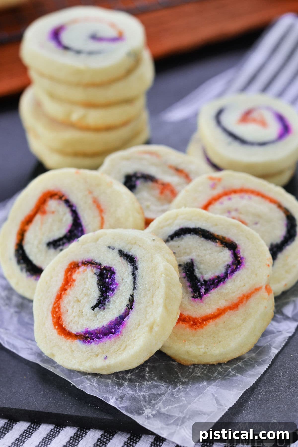 A beautifully arranged display of Spiral Halloween Cookies, made with vibrant orange, black, and purple sugar rolled into a captivating spiral design. A stack of cookies sits in the background, with five fanned out in front to highlight the neat swirls.