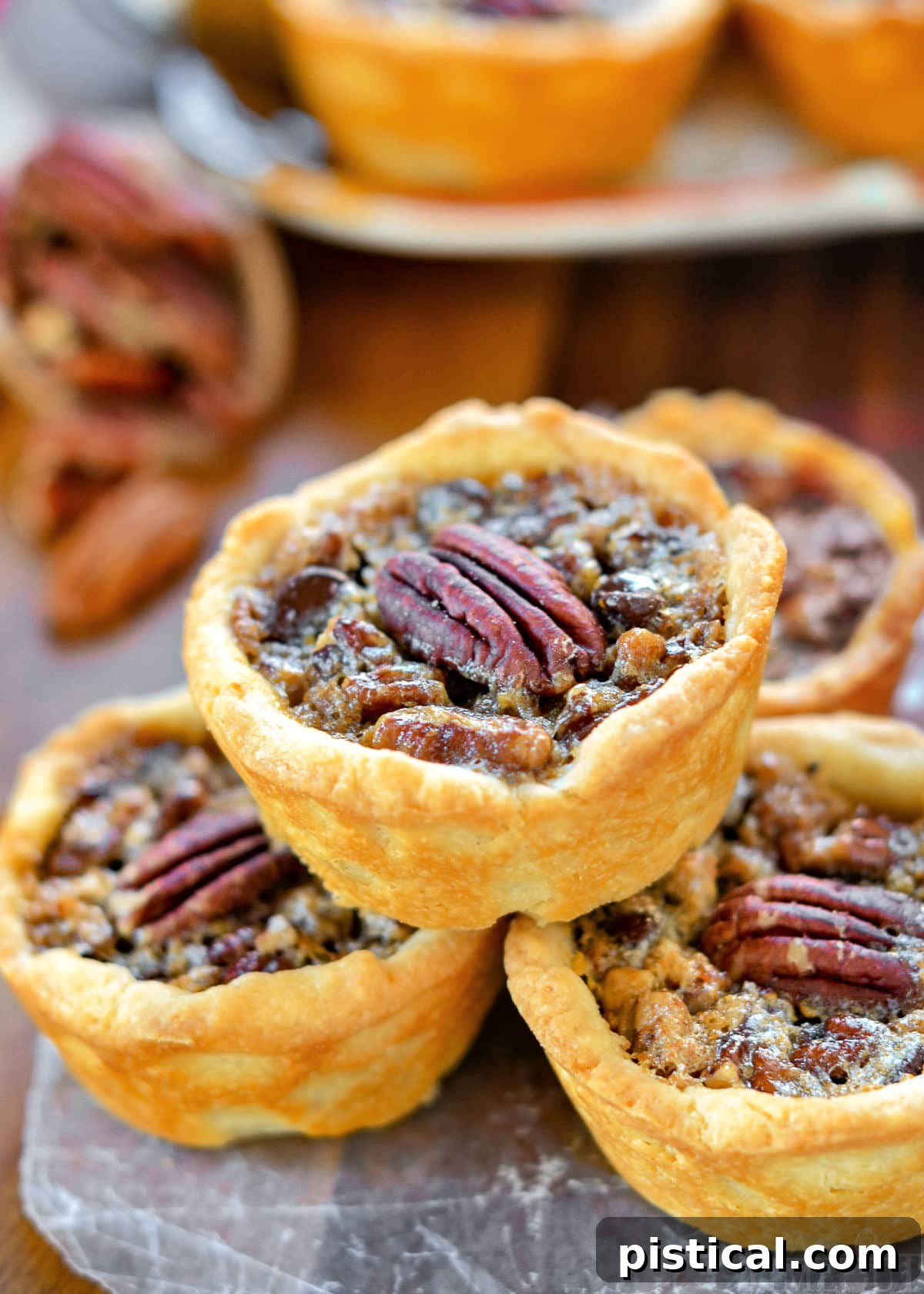 Four mini pecan pies elegantly stacked in a pyramid, with more pies artfully arranged in the background. Each mini pie is beautifully topped with a glistening pecan half, showcasing its inviting golden crust and rich filling.