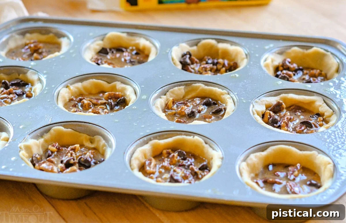 A muffin tin filled with unbaked mini pecan pies, each featuring a golden crust, rich filling, and a pecan half on top, ready to be baked to perfection.