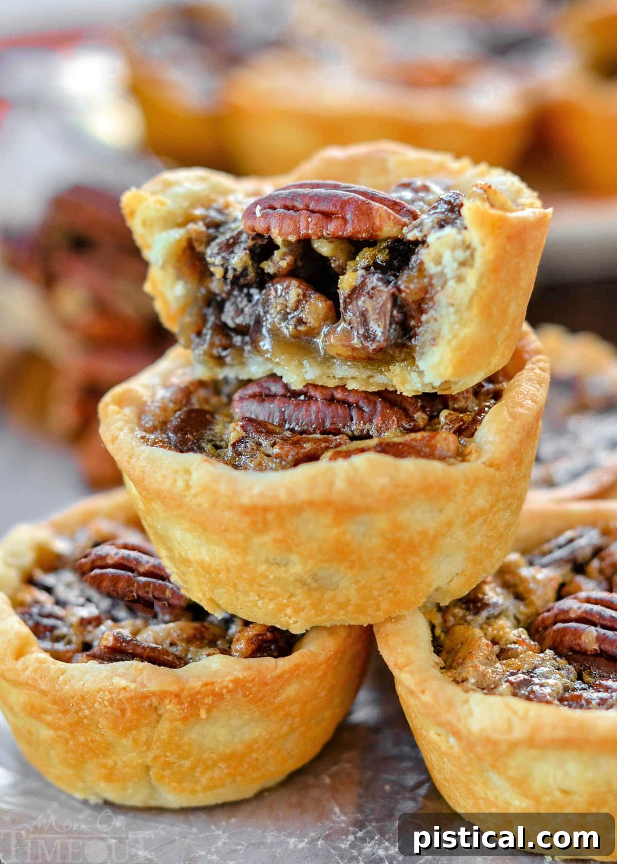 Five beautifully baked mini pecan pies stacked in a pyramid, with additional pies in the background. The top pie is cut in half, revealing the rich, set filling. Each pie is crowned with a whole pecan half, inviting a delightful taste.