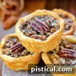 Four mini pecan pies stacked in a pyramid with more pies in the background. each mini pie is stopped with a pecan half.