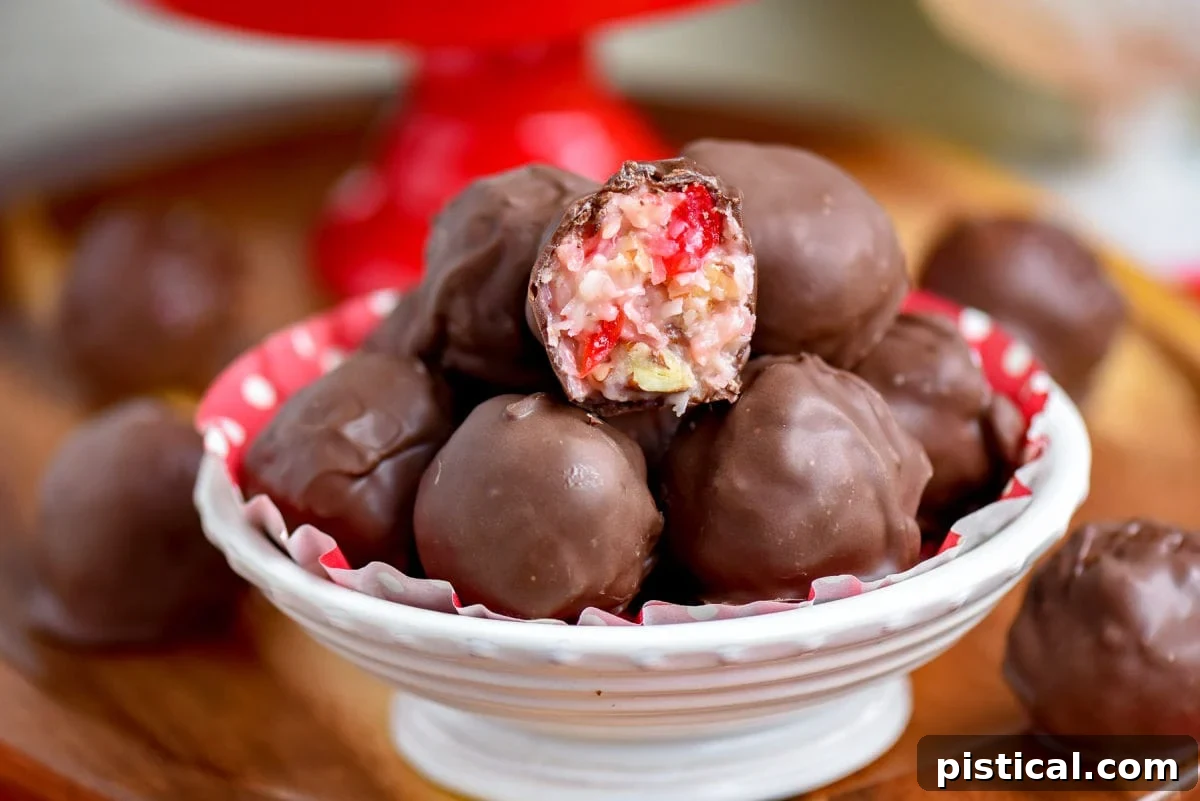 A white bowl brimming with Martha Washington candies. One candy is broken open to reveal its delightful coconut and cherry interior, with a festive red cake stand blurred in the background.