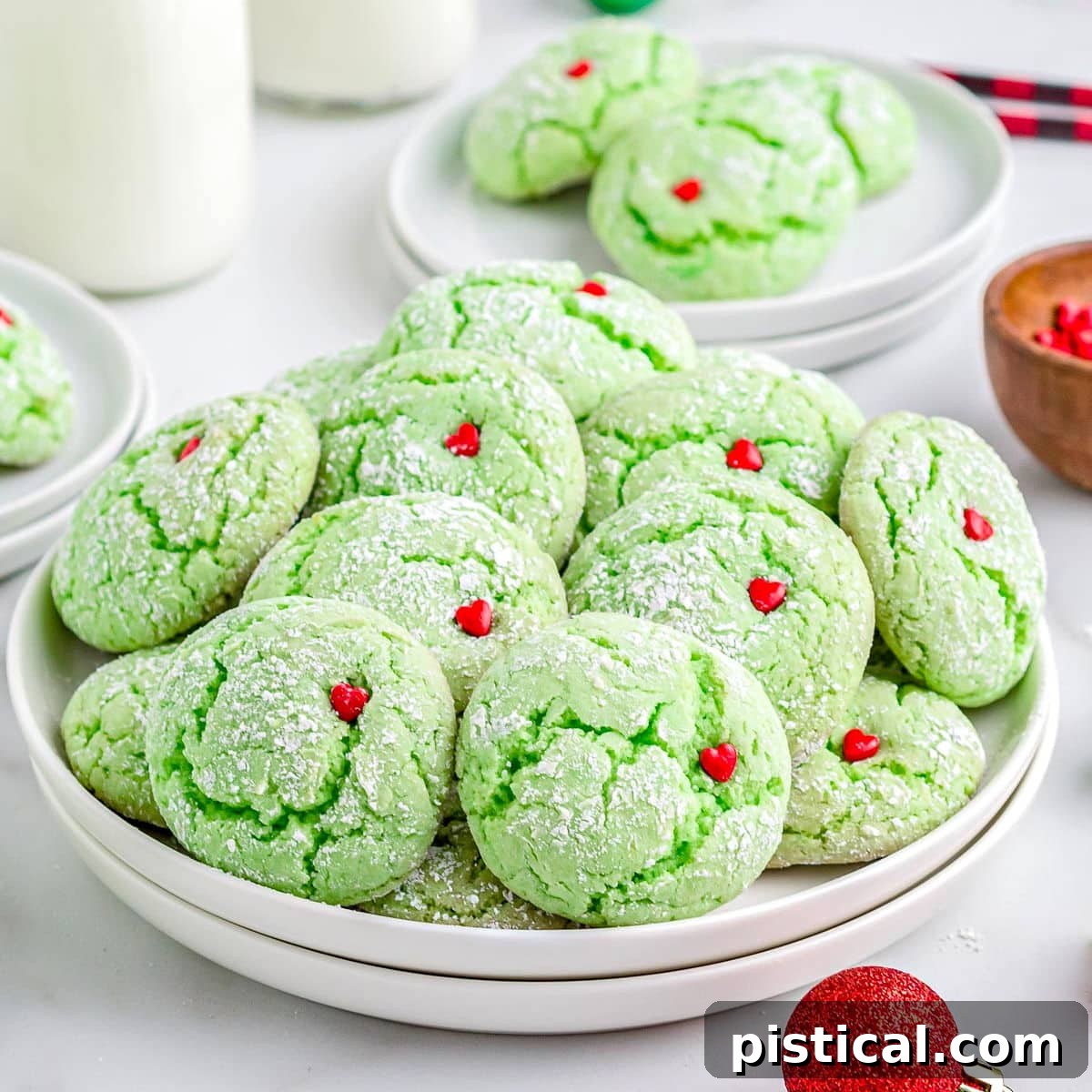 grinch cake mix cookies with red heart sprinkles sitting on white plate with milk jars in the background and Christmas ornaments scattered about.