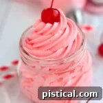 Smooth Maraschino Cherry Frosting piped into a clear jar, topped with a whole maraschino cherry for garnish.