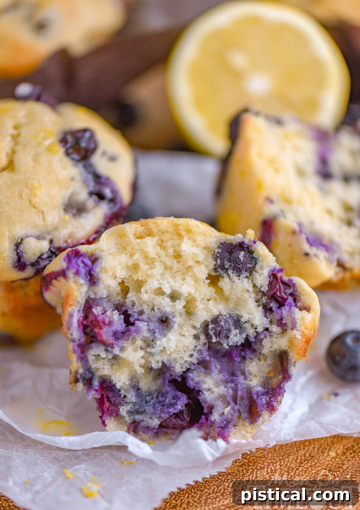 Zesty Lemon Blueberry Muffins with Cream Cheese Swirl 4 Blueberry Lemon Muffin split in half sitting on white parchment with more muffins and lemon halves in the background.