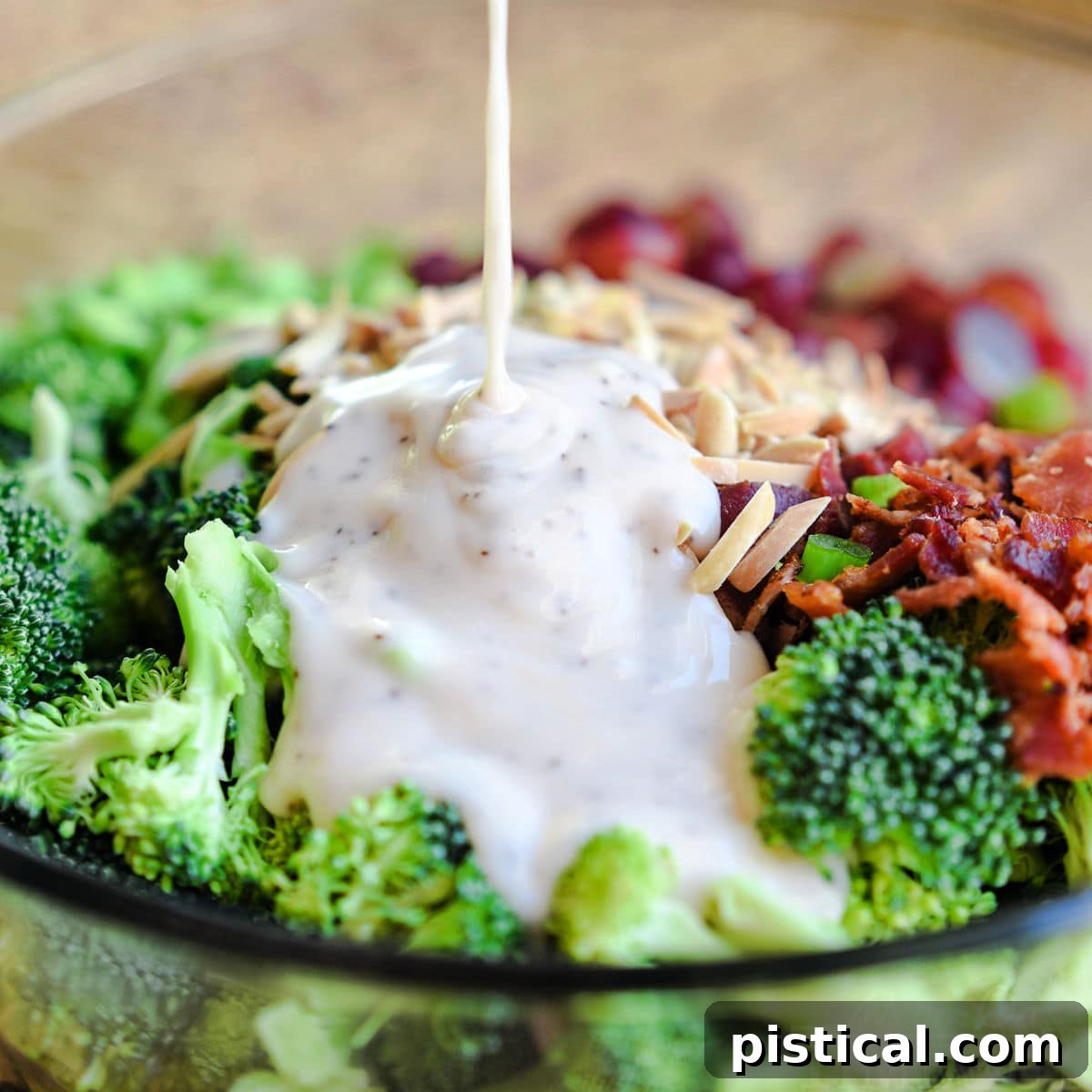 square image showing broccoli salad dressing being drizzled onto the rest of the ingredients in a large glass bowl.