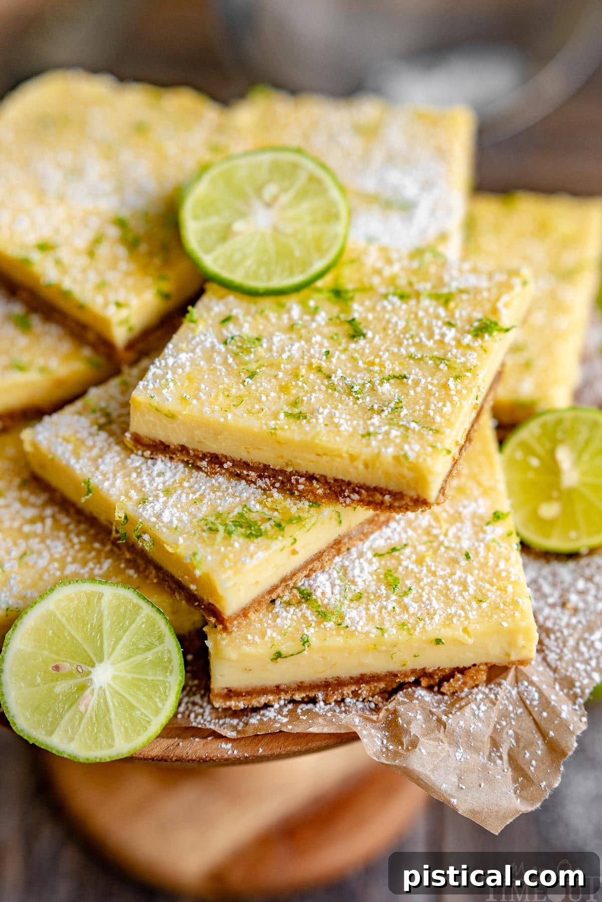 Zesty Key Lime Squares 6 A close-up shot of several delicious Key Lime Pie Bars piled high on a rustic wood cake stand, lined with parchment paper. The bars are beautifully garnished with freshly grated lemon and lime zest, and a generous dusting of powdered sugar, emphasizing their vibrant and inviting appearance.