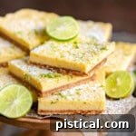wood cake stand piled high with key lime bars topped with key lime zest and a light dusting of powdered sugar. Slices of lime are scattered around the bars.