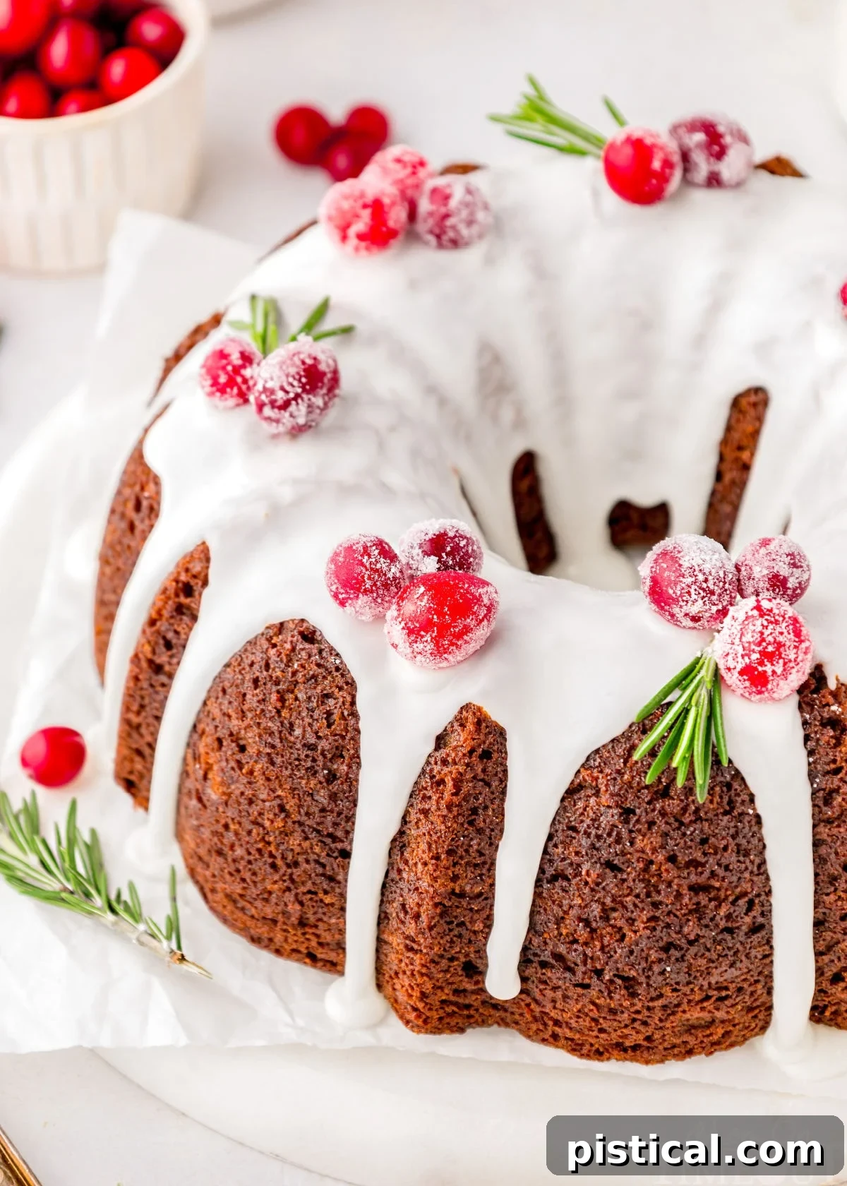 Cozy Gingerbread Bundt Delight 2 Gingerbread bundt cake topped with vanilla icing and cranberry and rosemary garnishes. The cake is on a white cake stand, adorned with festive decor.
