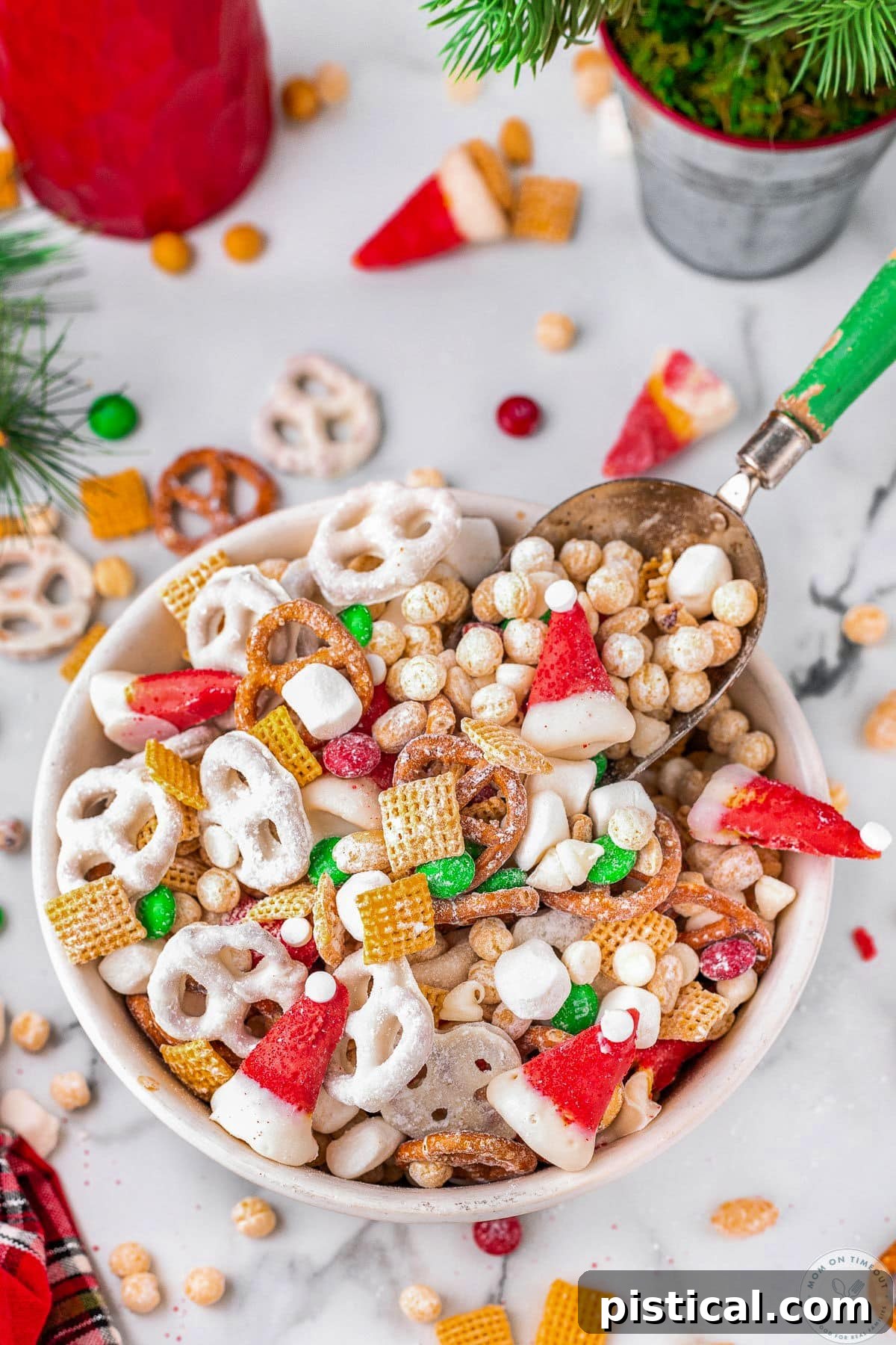 A white bowl brimming with Christmas snack mix, complete with a small scoop. Pretzels, peanuts, and various candies are artfully scattered around the bowl, highlighting the mix's diverse components.