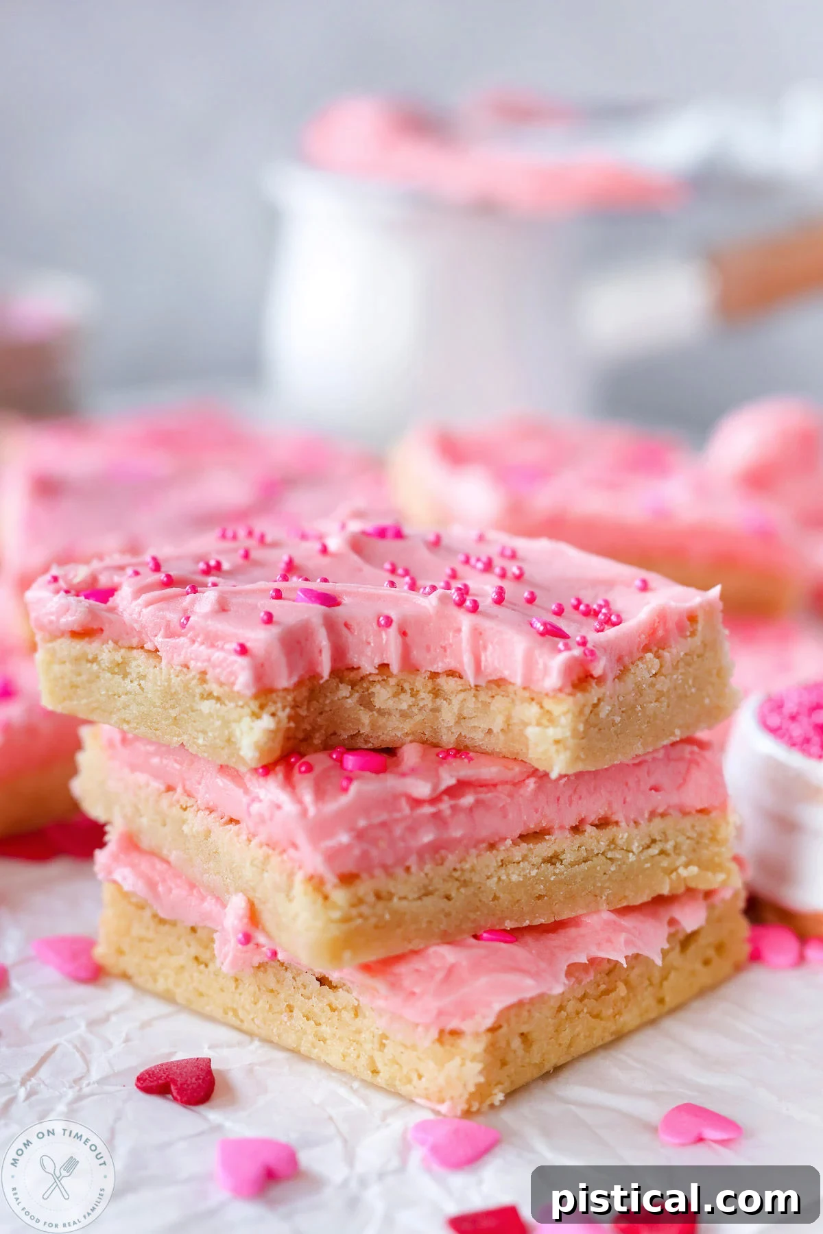 Three sugar cookie bars frosted with pink cherry frosting stacked on white parchment paper. Top bar has a bite taken out of it.