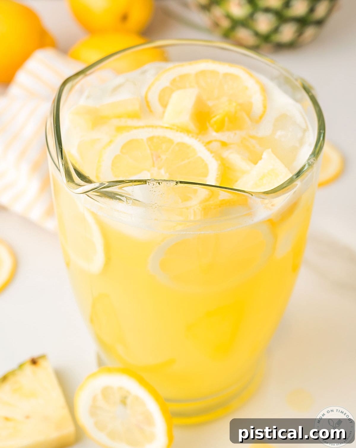 Angled overhead shot of pineapple lemonade recipe in glass pitcher garnished with lemon slices and pineapple chunks.